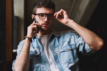 Portrait of serious hipster guy in trendy eyeglasses calling to customer service from cellular...