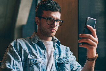 Concentrated pensive hipster guy holding smartphone and reading social news, young caucasian man in...