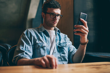 Concentrated pensive hipster guy holding smartphone and reading social news, young caucasian man in...