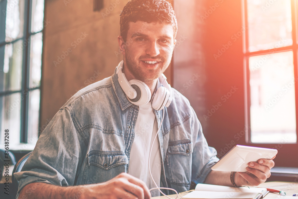 Poster portrait of cheerful hipster guy with touch pad in hand looking at camera while installing media app