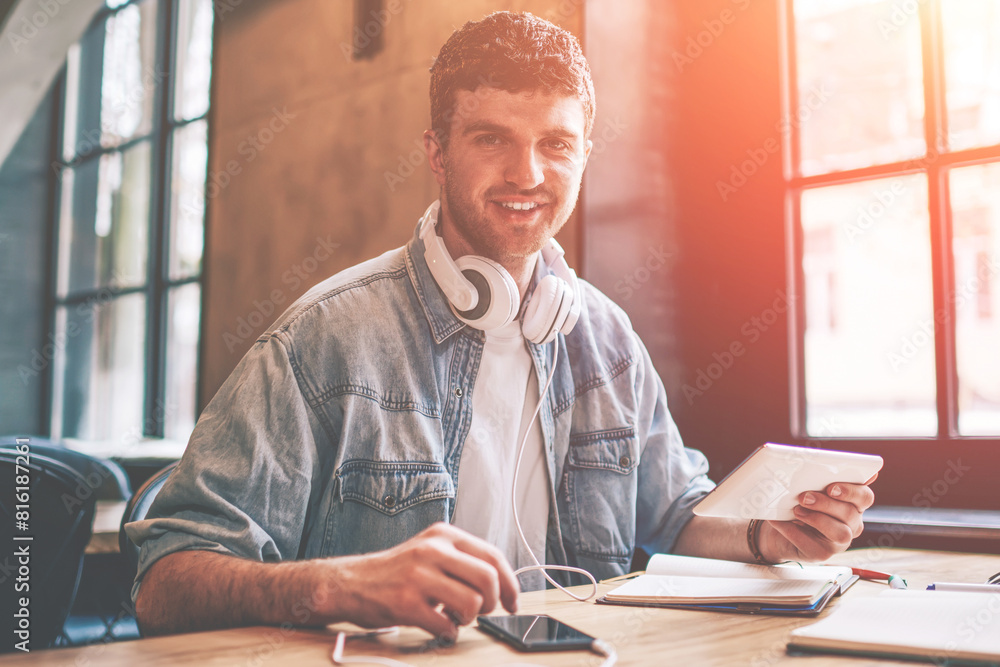 Poster portrait of cheerful hipster guy with touch pad in hand looking at camera while installing media app