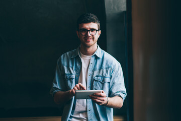 Portrait of cheerful hipster guy with cute smile on face using digital tablet for searching...