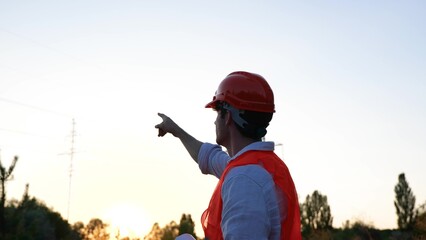 Happy male engineer planning new project while pointing to the high voltage power lines. Highest voltage transmission. Energy industry concept. Slow motion