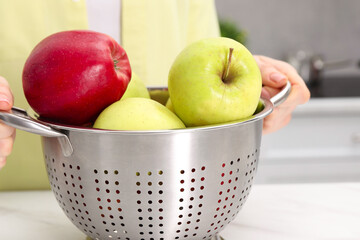 Woman holding colander with fresh apples at table indoors, closeup
