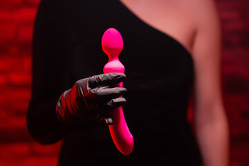 Pink stylish vibrator for masturbation in the hands of a girl wearing black gloves, dressed in a black dress on a red background. Massager with multiple speeds. Sex shop products, adult gifts