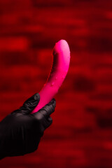 Pink stylish vibrator for masturbation in the hands of a girl in black gloves on a red background. Massager with multiple speeds. Products for sex shop, gifts for adults