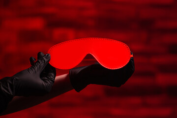 Close-up of female hands in black latex gloves holding a red BDSM headband on a red background. BDSM - outfit for adult sex games