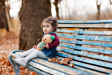 Child's thoughtful face in autumn park, sitting on bench in and holding her teddy
