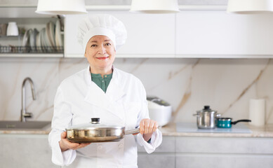 Experienced hostess in chef hat and white cook uniform is standing in comfortable modern kitchen...