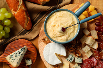 Fondue pot with tasty melted cheese, fork and different snacks on wooden table, top view