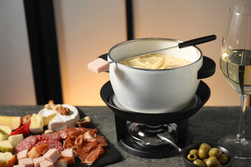 Fork with piece of ham, fondue pot with melted cheese, wine and snacks on grey table, closeup