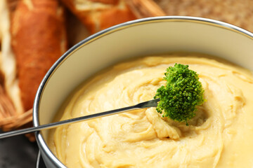Dipping piece of broccoli into fondue pot with melted cheese on blurred background, closeup