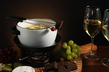 Forks with pieces of grape, bread, fondue pot with melted cheese, wine and snacks on table, closeup