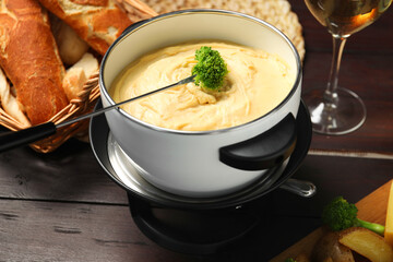 Fork with piece of broccoli, melted cheese in fondue pot and snacks on wooden table, closeup