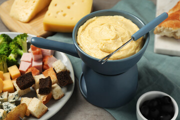 Tasty melted cheese in fondue pot, fork and snacks on grey table, closeup