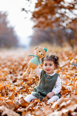 Playful autumn moments. Sweet little girl sitting among autumn leaves, showing her teddy and smiling