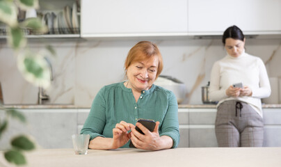 Happy mature mother and her daughter browsing social media on phones in kitchen