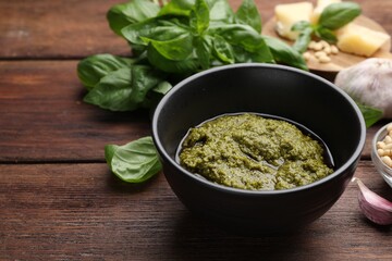 Tasty pesto sauce in bowl, basil, garlic and pine nuts on wooden table, closeup