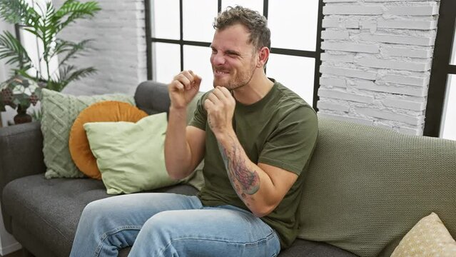Joyful young man sitting on sofa at home, victoriously throwing winner gesture, excitedly screaming in triumph of success, while smiling broadly.