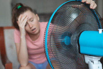 Young woman using electric fan at home in living room, sitting on couch cooling off during hot weather, suffering from heat, high temperature