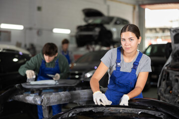 Young female auto mechanic in blue overalls preparing car for painting, sanding removed bumper with...