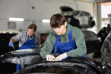 Young guy mechanic in uniform polishes part of car with sponge in car service station