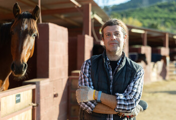 Adult male stables worker in plaid shirt holding brushes and gloves before cleaning horses from...