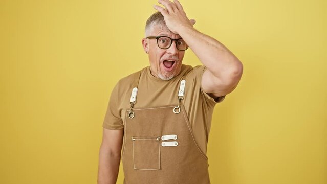 Oops! middle age, grey-haired man, standing in an apron and glasses, hand on head in foolish regret. forgot something critical? memory fail on isolated yellow background. annoyed with his blunder.