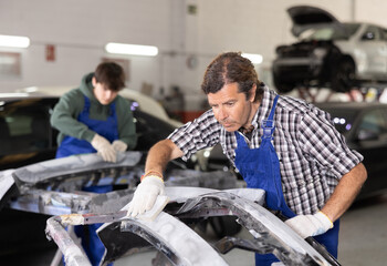 Experienced auto body technician in blue overalls focused on smoothing surface of car parts using...