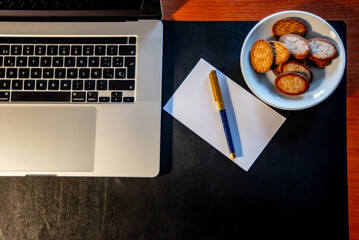 Working desk with a notebook, a red cup of coffee, some sweet cookies on a plate, and a blue and gold fountain pen, on a sheet of white paper. The background is light wood.