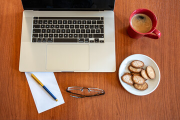 Working desk with a notebook, a red cup of coffee, some sweet cookies on a plate, a pair of...