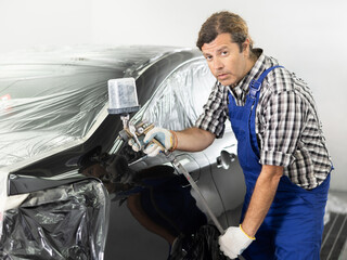 Focused middle aged man in casual checkered shirt and blue overalls working in car painting...