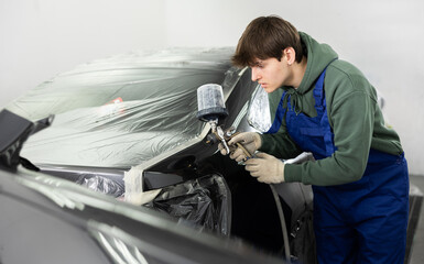 Focused young guy in casual olive hoodie and blue overalls working in car painting workshop, using...