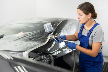 Professional young female auto painter in blue overalls working in automotive workshop, focused on...