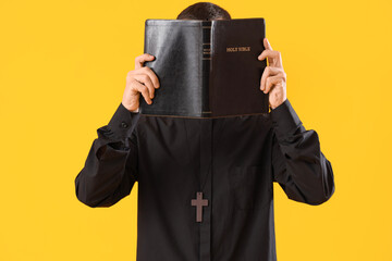 Young priest with Holy Bible on yellow background