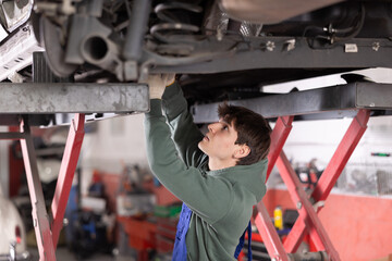 Young guy mechanic in uniform repairs underbody of car in car service station