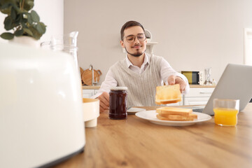 Young man with laptop having tasty jam toasts for breakfast in kitchen
