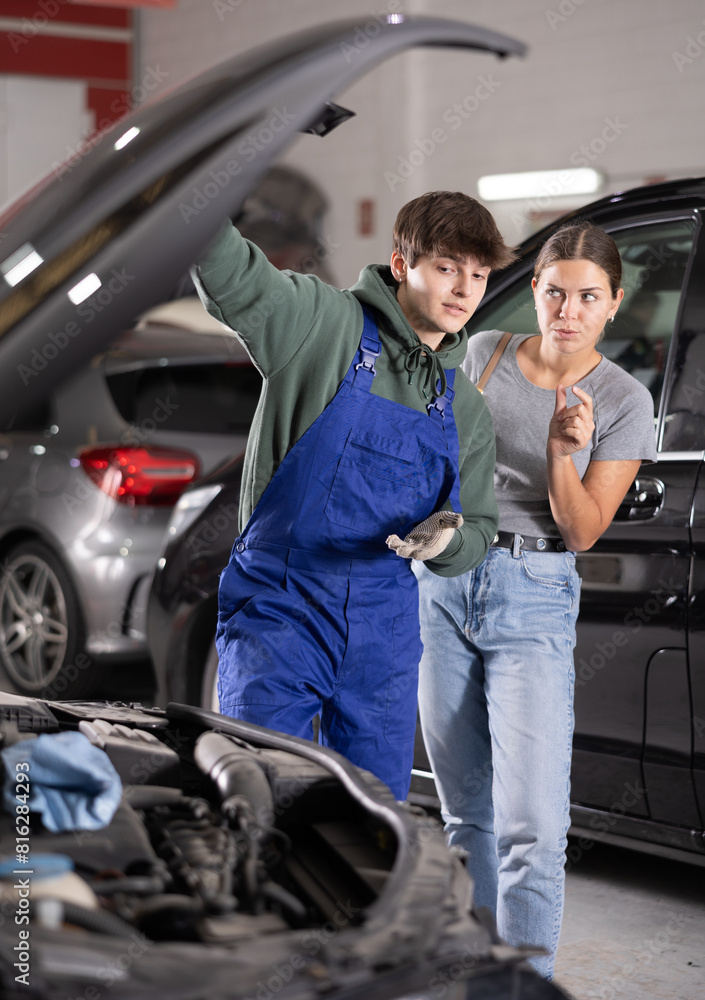 Wall mural Young guy mechanic advises young woman client on repairing under hood of car in car service station