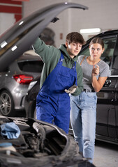 Young guy mechanic advises young woman client on repairing under hood of car in car service station