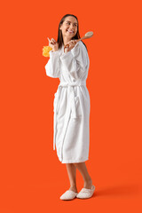 Young woman in bathrobe with loofah and massage brush on orange background