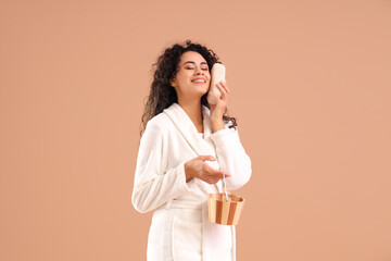 Young African-American woman in bathrobe with loofah and bucket on brown background