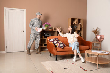 Man in military uniform greeting his wife with flowers and heart-shaped gift box at home....