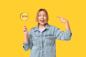 Surprised adult woman pointing at speech bubble with word WOW on yellow background