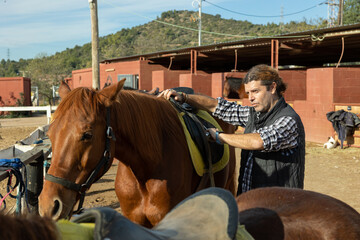 Portrait of experienced adult stables worker in plaid shirt standing close to saddled horse in...