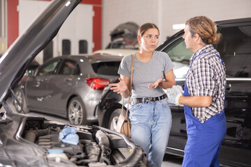 Adult male mechanic advises young woman client on repairing under hood of car in car service station