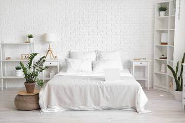 Interior of light bedroom with houseplant and laptop on white bed