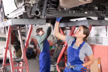 Confident young female auto mechanic in blue overalls standing under car on lift in service...