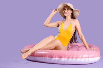 Beautiful woman in swimming suit and hat sitting on inflatable mattress against purple background
