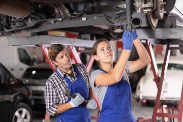 Two focused professional mechanics, middle aged man and young woman, wearing blue overalls,...
