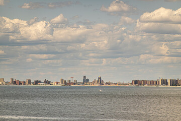 Fototapeta premium New York, NY, USA - August 1, 2023: Maimonides Park on Coney Island beach in wide landscape under blue cloudscape. Gray ocean water up front. Brown buildings belt